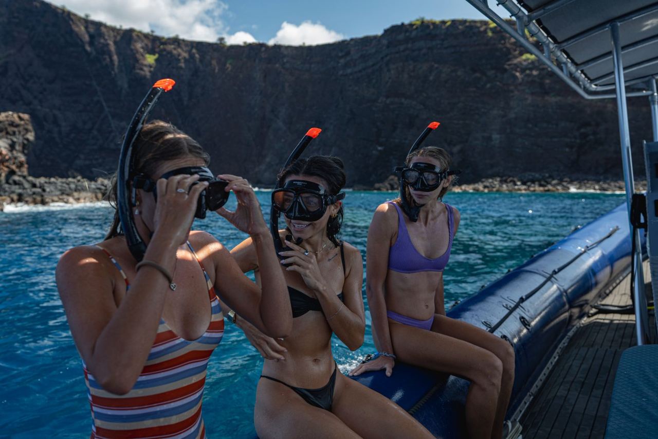 Three guests in masks sitting on the raft tube against the cliffs of Lānaʻi
