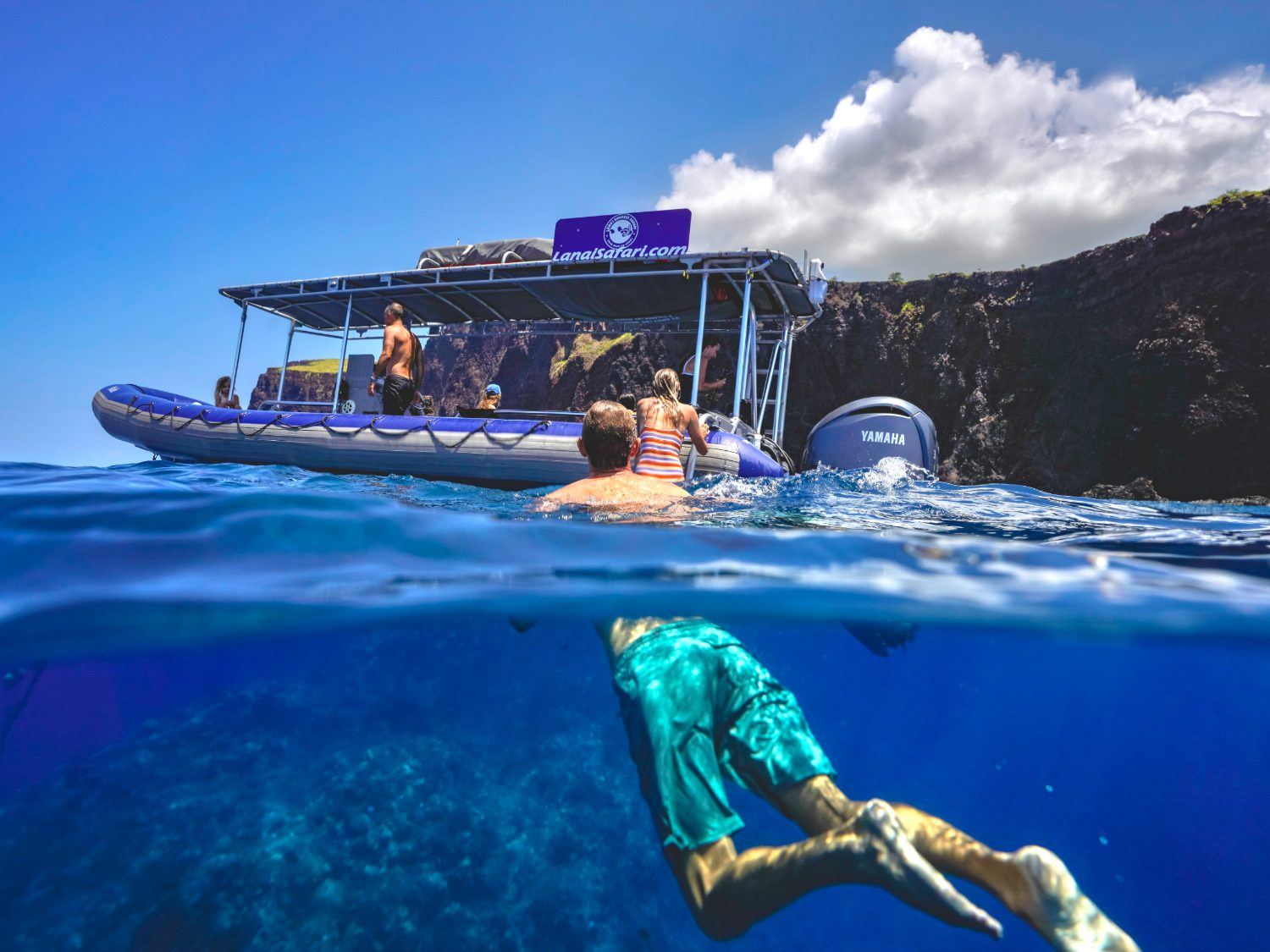 A split over-under view: a snorkeller underwater in the foreground with the raft and the Lānaʻi cliffs above the surface