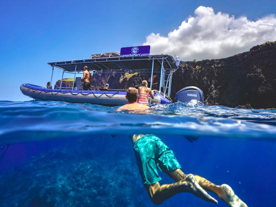 A split over-under view: a snorkeller underwater in the foreground with the raft and the Lānaʻi cliffs above the surface
